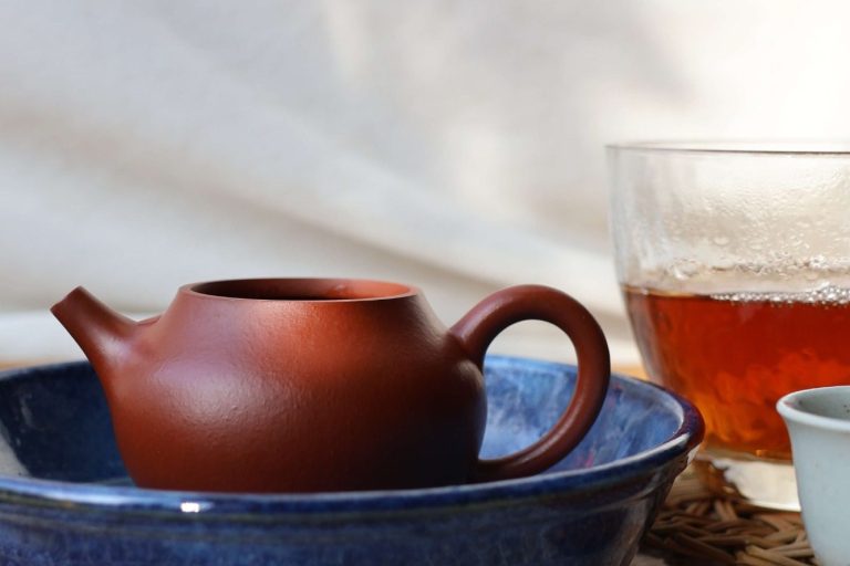 A red clay teapot in a blue ceramic tray, with a glass pitcher containing dark red-brown tea liquor and porcelain cup in the background