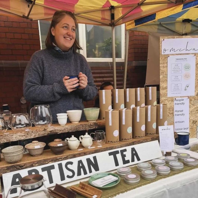 Meg stands behind her market stall featuring teas and handcrafted teaware, holding a bowl of tea and smiling at a customer out of frame