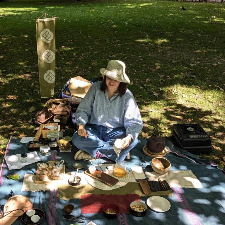 Meg sits on a blanket in the grass in dappled sunlight, preparing tea