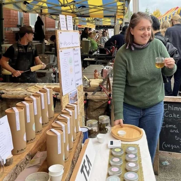 Meg stands next to her table at the Tobacco Factory market with a glass pitcher of tea in her hand