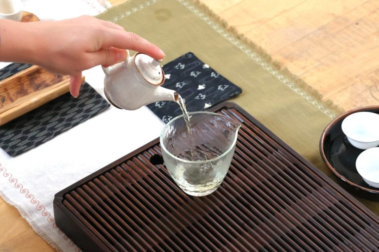 Meg sits at a small table with a tray containing a small teapot and cups, with tea fields in the background
