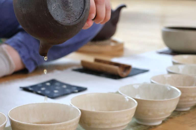 Meg sits at a small table with a tray containing a small teapot and cups, with tea fields in the background