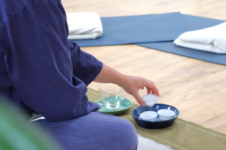Tea being poured into a small white porcelain teacup on a bamboo coaster, with light falling across the cup and the rest of the table in shadow